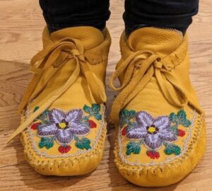 A pair of beaded mocassins is worn by someone. Only their feet show.